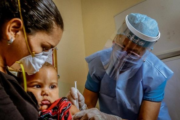 1歳のマルセロくんに定期予防接種のワクチンを接種する、保健員。新型コロナウイルス感染症の感染予防のため、ユニセフ支援物資の個人用防護具を身につけている。（ベネズエラ、2020年7月）