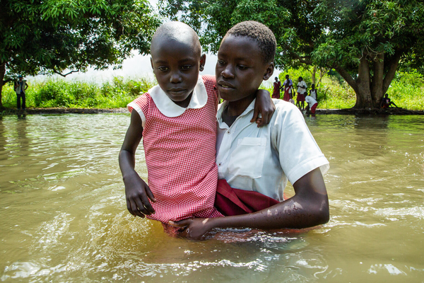 洪水で腰まで水位が上がった道を、下級生の女の子を抱きながら歩く上級生の女の子。(南スーダン、2021年9月撮影)