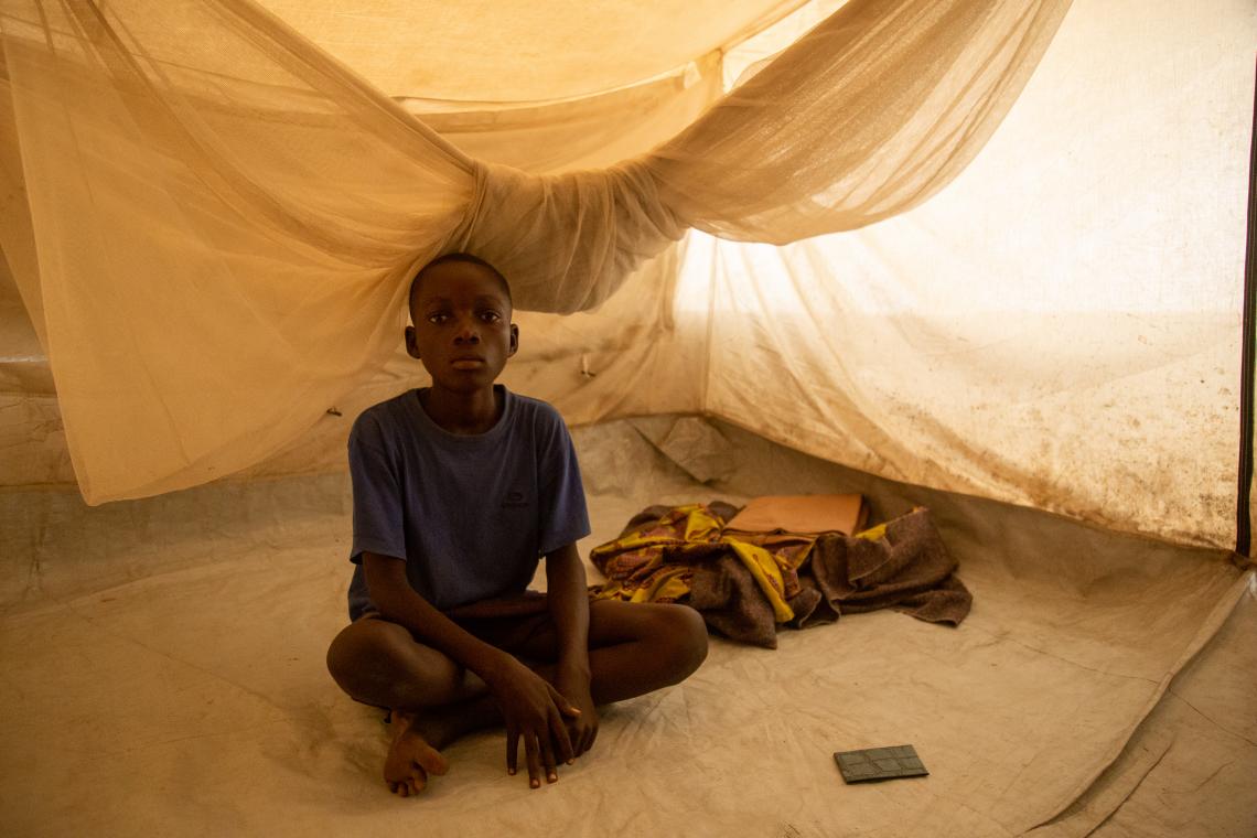 家族と共有しているテントの中に座っているエマニュエルくん（15歳）。そばにあるのは、家族が唯一持っている毛布。母親のマリーさんは、「テントでの生活は大変です。日中は暑くて息苦しく、夜はとても寒いのです」