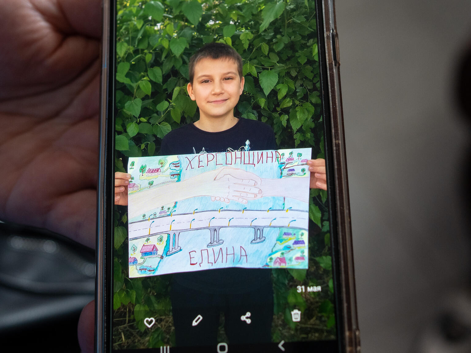 「Kherson is united（ヘルソンは団結している）」というメッセージを描いた絵を見せるイリヤの写真が、スマートフォンで映し出されている。