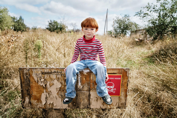 「地雷の危険」と書かれた看板の上に座る6歳のレオニードさん。戦闘が激化しても家族はハルキウ州の自宅に留まり続けた。地元の学校は破壊されてしまったため、レオニードさんはオンラインで授業を受けている。(ウクライナ、2023年9月撮影)