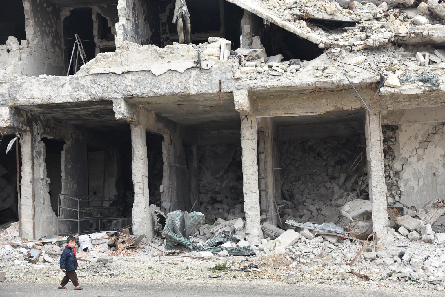 戦闘により破壊された建物の傍を歩く男の子。彼のように、平和なシリアを知らない子どもが多くいます。