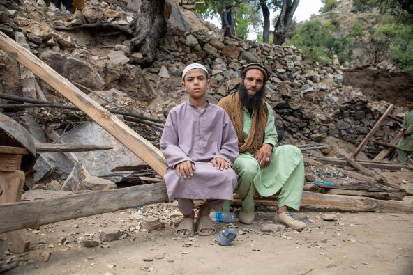 クナール県で、地震により倒壊した自宅前で座り込む親子。母親ときょうだいの一人が亡くなった(アフガニスタン、2025年9月3日撮影)
