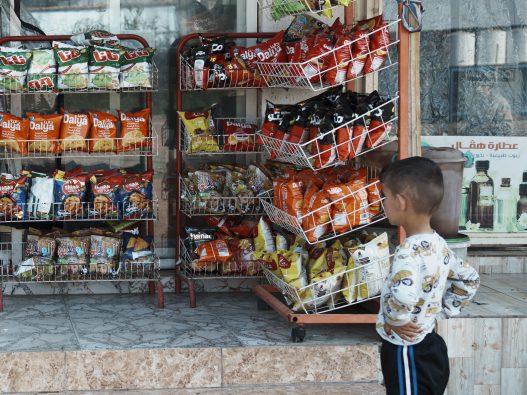 エルビルの難民キャンプ内にある商店の前で、陳列されているポテトチップスの袋を見つめる男の子（イラク、2024年3月5日撮影）