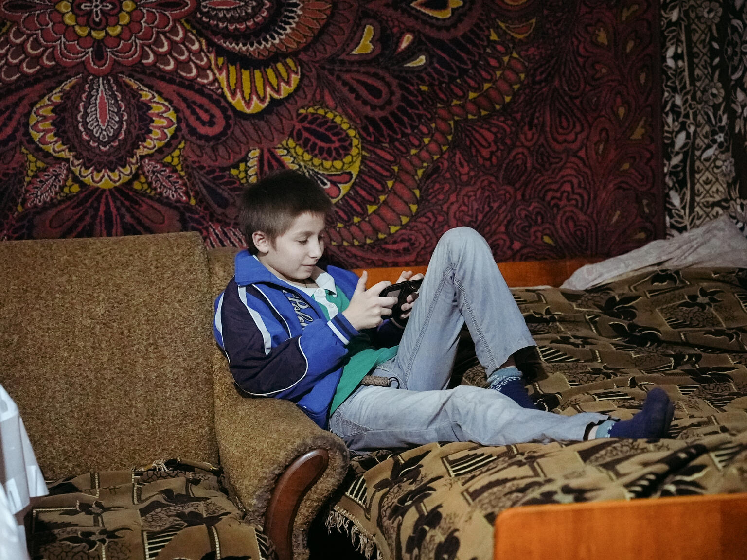 自宅のリビングで、スマートフォンを使って遊ぶ男の子。戦争下で停電が続くなかでも、こうした何気ない日常を保つことが子どもにとってとても重要である（ウクライナ、2025年11月6日撮影）