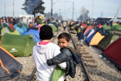 線路の周りに立てられたテントの間を、弟を抱いて歩く難民の少年。（ギリシャ）