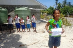 新しく設置された雨水貯水備蓄施設で安全な飲み水を手にすることができるようになったキリバスの子どもたち。「水は、飲み物」と書かれた紙を手に笑顔を見せる少女。