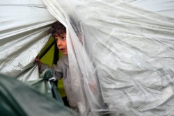 一時的な住居となっている、ビニールシートで作られたテントから、顔をのぞかせる男の子（ギリシャ）