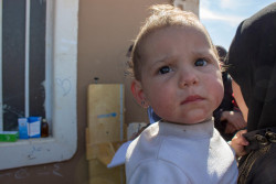On March 17 2018 in the Syrian Arab Republic, a woman carrying a child waits with other people outside a building at the Herjeleh collective shelter in Eastern Ghouta. The shelter is among three in Rural Damascus housing thousands of people displaced from their hometowns and villages in East Ghouta by intense fighting. All of the shelters are well over capacity, and more people arriving each day. Water and sanitation facilities, non-food items such as blankets, mattresses, clothing and other essential items are all needed urgently. UNICEF and partners are providing water, sanitation and hygiene (WASH), health, nutrition, child protection and other support. UNICEF is supporting water trucking and the provision of water tanks to ensure access to safe water, and is distributing bottled water, high-energy biscuits and other water, and health and nutrition supplies. UNICEF-supported mobile health teams are also providing basic medical assistance and vaccinations for children and mothers.