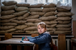 Lera Nagormay, 10, sits for a photograph in a classroom at school in Marinka, Donetsk Oblast, Ukraine, Wednesday 22 November 2017. When conflict broke out in 2013, Marinka was heavily contested. "One time, when I walked to school" Lera says, “when I arrived all the kids were already in the shelter. Shelling had started while I was on my way, and I had to rush in there.  The school still has weekly drills, corralling students into the bunker beneath the building. In a long cold room, seats line one wall for the smaller children.  "Sometimes we bring our toys down here so we don't get bored waiting," Lera says. Today, a police officer wearing camouflage and armed with a rifle stands guard in the lobby of the school. After one child was shot in the arm in the playground, children are not permitted to play outside. Field trips are out of the question due to the risk posed by mines and unexploded ordnance. "We have to stay inside the school all the time, and are not allowed to play outside during the breaks. The boys play football in the school's corridors" Lera says, "they can't break the windows because of the sandbags." As of December 2017, the situation in eastern Ukraine remains volatile, and violence continues despite the latest ceasefire agreements committed on 19 July 2017. The lives of children and their families, especially those living along the contact line continue to be at risk. According to a December 2017 report by the United Nations Office for the Coordination of Humanitarian Affairs, millions of people are continuing to suffer unnecessarily due to the entrenched political impasse and the ongoing armed conflict. Despite many attempts at a ceasefire, hostilities continue with almost daily shelling, frequent localized clashes, and rapidly escalating mine and unexploded ordinance contamination.  The conflict has taken a severe toll on the education system, affecting students, teachers, administration and education facilities, hundreds of which hav