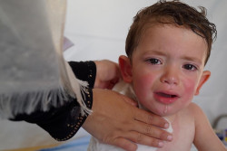 On 6 July 2018 in Jordan, Omar, 2 years, from Dera’a in Syria, in the UN-supported health clinic at the Jaber Nasib crossing point. His mother brought him to the clinic because he has a fever. The family are from Dera’a.  On 19 June 2018, an escalation of violence and fighting erupted in the southwestern Syrian governate of Dera’a resulting in the displacement of at least 270,000 people. Of these, an estimated 60,000 people fled to the Nasib-Jaber border crossing between Syria and Jordan where they are living in dire humanitarian conditions in the Free Zone – a no man’s land on the Syrian side of the border. The majority are women and children. The UN has warned that without urgent assistance to meet families’ needs, the situation could quickly deteriorate into a humanitarian catastrophe. In coordination with the Government of Jordan, the UN has been assisting families in the Free Zone with trucks of humanitarian supplies and staff deployed within 48 hours of the first arrivals on 28 June. UNICEF Jordan deployed staff and supplies, as part of the UN response, and has been operating daily to deliver urgent humanitarian assistance to women and children. Children, forced to flee horrific violence, have arrived to the border area terrified, injured and, in some cases, separated from their parents. Mothers have reported that their children are crying, clingy and scared by any loud noises. Women and children are struggling to survive and stay healthy without toilets or clean water. Children forced to live out in the open with little or no shelter are suffering severe dehydration as temperatures climb to 34ºC. UNICEF is leading the WASH response in the service area and inside the Free Zone. This assistance has included: delivery of 15 trucks of urgent WASH supplies containing jerry cans, soap and hygiene kits to the border for distribution; constructed latrines, water stands with drinking water and built a septic tank for the service area clinics and humani