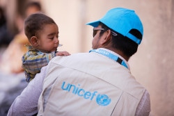 On 20 March 2018 in Adra in eastern Ghouta in the Syrian Arab Republic, a UNICEF Syria child protection specialist holds 6-months-old baby Yusuf as his mother queued for services. Since 11 March 2018, more the 50,000 people have left Eastern Ghouta and thousands more are reported to be on exit points awaiting transportation to collective Shelters. In Adra, an estimated 5,400 people are currently staying in three schools-turned-shelters in the area, while over 13,000 have sought shelter in an abandoned institute, formerly used for electrical studies.  At the shelters, children and their families are living in the most basic conditions. Overcrowding and lack of existing water and sanitation systems are increasing families’ hardship. Many are forced to sleep out in the open, bearing the day’s scorching sun and the night’s dropping temperatures. Families have arrived to the shelters with very few personal belongings. UNICEF and partners were there to support in the four Adra shelters since families started arriving last week in what was the largest displacement from one location in one day, since the beginning of the Syrian conflict. Emergency water trucking is ongoing in all four shelters into water tanks installed by UNICEF. UNICEF has also installed pre-fabricated latrines at the three schools and is finalizing the installation of toilets and showers at the electricity compound. However, due to the huge influx of people on daily basis, sanitation remains a challenge. Three mobile health teams are providing primary healthcare services for children and mothers, including the provision of nutritional supplements to prevent and treat malnutrition as well as much-needed vaccinations. Most shelters do not have the capacity to accommodate the large number of people arriving. Some sites are not suitable to host IDPs and overcrowding is impeding needed shelter and WASH rehabilitation. As a result many families are staying out in the open and there are insufficient WA