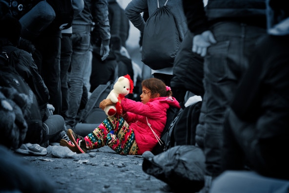 [A CHILD IS A CHILD CAMPAIGN PHOTO]  A little girl is sitting on the ground playing with her teddy bear, while people around her wait to enter the Vinojug reception center near the town of Gevgelija, in the former Yugoslav Republic of Macedonia.