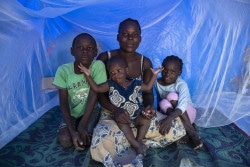 22 April 2019. Lorenza Sulemane (23) sits with her baby Sulemane Childo (10 months old) and her son, Yezezu Childo (8) and her daughter Enesh Childo (6) under their mosquito net in a tent at the Picoco Accommodation Centre in Beira, Mozambique. She shares the tent with six people. The are from the Praia Nova Area in Beira and their home got destroyed during Cyclone Idai. They found temporary accommodation at the Agostino Neto School and were then moved to the Picoco Accommodation Centre.  She says that she uses the mosquito net to sleep under but that she does not know the reasons why she has to sleep under the mosquito net. On the 15th of March 2019, a cyclone ripped through central Mozambique. Heavy rains caused the banks of the Buzi river to break creating an ‘inland ocean.’ Floods tore through communities, destroying everything in their path. Thousands of standard families, many of them with young children, sought safety on top of high buildings waiting to be rescued.  Flooding has left pools of stagnant water around communities so humanitarian responders are bracing for a surge in Malaria cases, in the last two weeks alone, there have been over 12,000 cases of malaria reported across the disaster area.