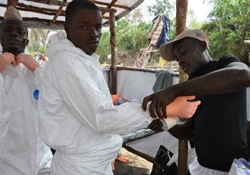 遺体をトラックから遺体安置所のテントまで搬送する準備として、
埋葬チームのメンバーが防護衣類の着用を手伝ってもらっている。